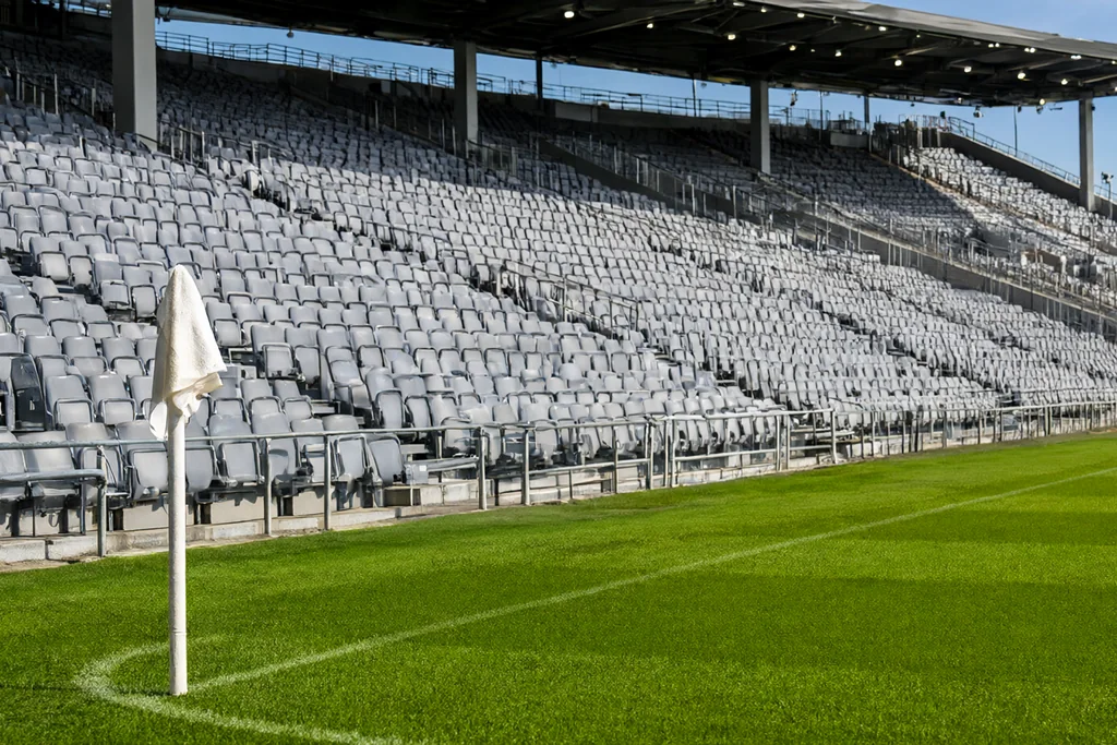 Grey plastic seats of an empty stadium stand above a green pitch with a corner flag